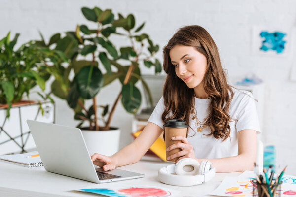 smiling young woman using laptop and holding paper cup while sitting at desk near headphones