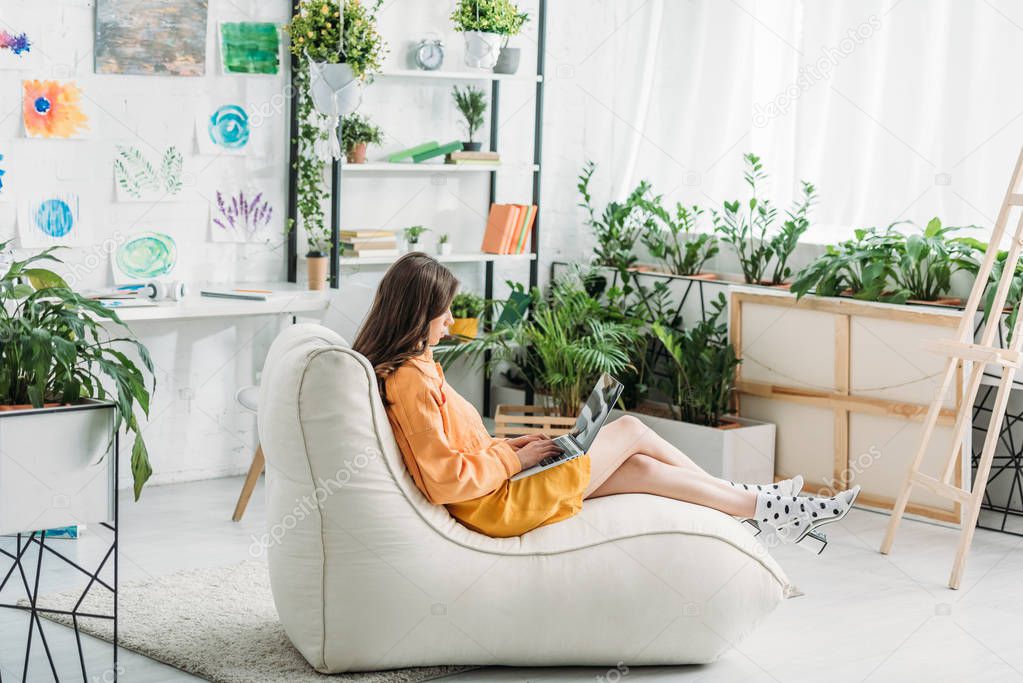 young woman using laptop while sitting on soft chaise lounge in light spacious room 