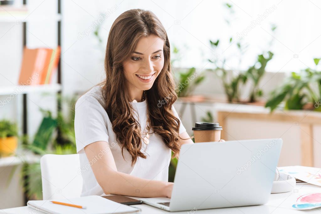 cheerful young woman using laptop and holding paper cup while sitting at desk at home
