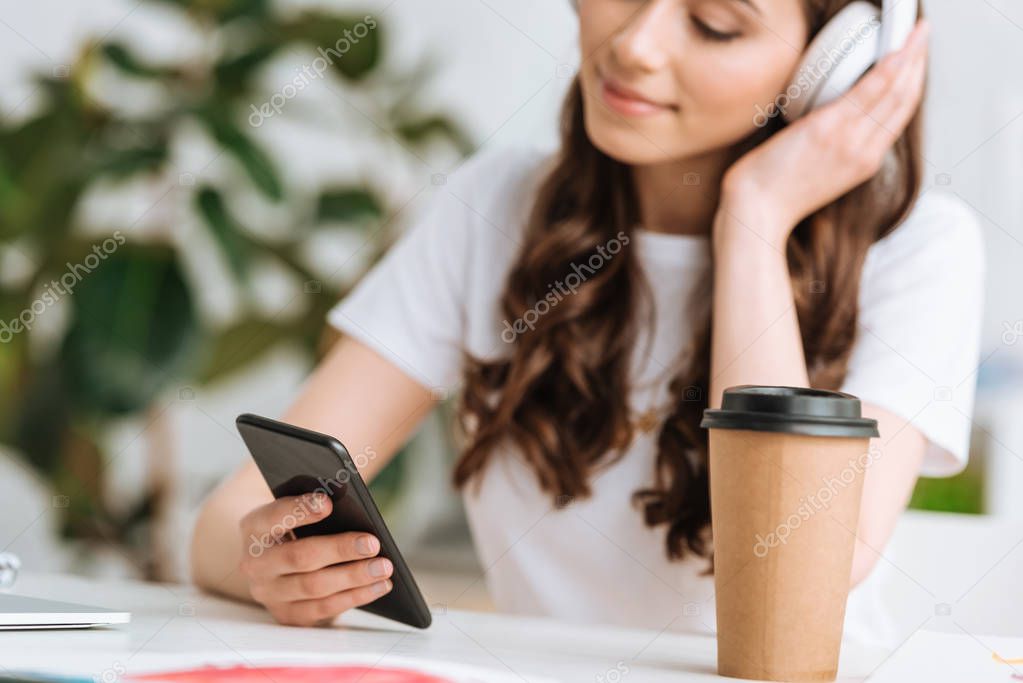 selective focus of young woman in headphones using smartphone while sitting near disposable cup
