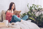 selektiver Fokus einer fokussierten jungen Frau, die Buch liest, während sie mit gekreuzten Beinen in der Nähe grüner Pflanzen zu Hause sitzt