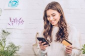 schöne junge Frau lächelt, während sie Kreditkarte hält und Smartphone benutzt