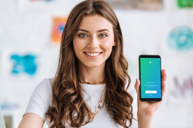 KYIV, UKRAINE - APRIL 17, 2019: Cheerful young woman looking at camera and showing smartphone with Twitter app on screen. clipart