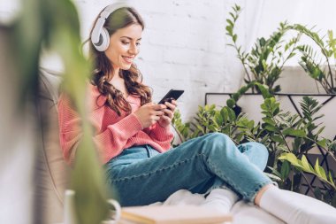 yeşil bitkiler le çevrili otururken akıllı telefon kullanarak gülümseyen kız seçici odak ve kulaklık müzik dinleme