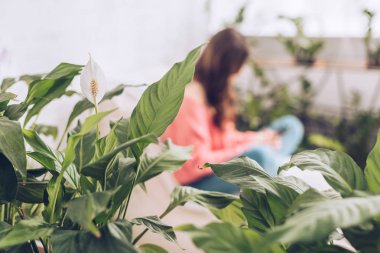 selective focus of young girl sitting in room with lush green plants clipart