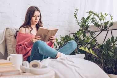evde yeşil bitkilerin yakınında çapraz bacaklar ile otururken kitap okuma odaklı genç kadın seçici odak