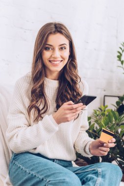 kredi kartı ve akıllı telefon tutarken kameraya bakan mutlu genç kadın