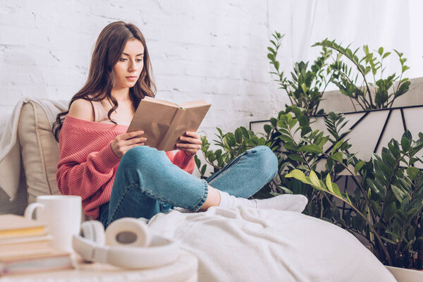 selective focus of focused young woman reading book while sitting with crossed legs near green plants at home