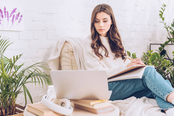 beautiful young woman writing in notebook and using laptop at home