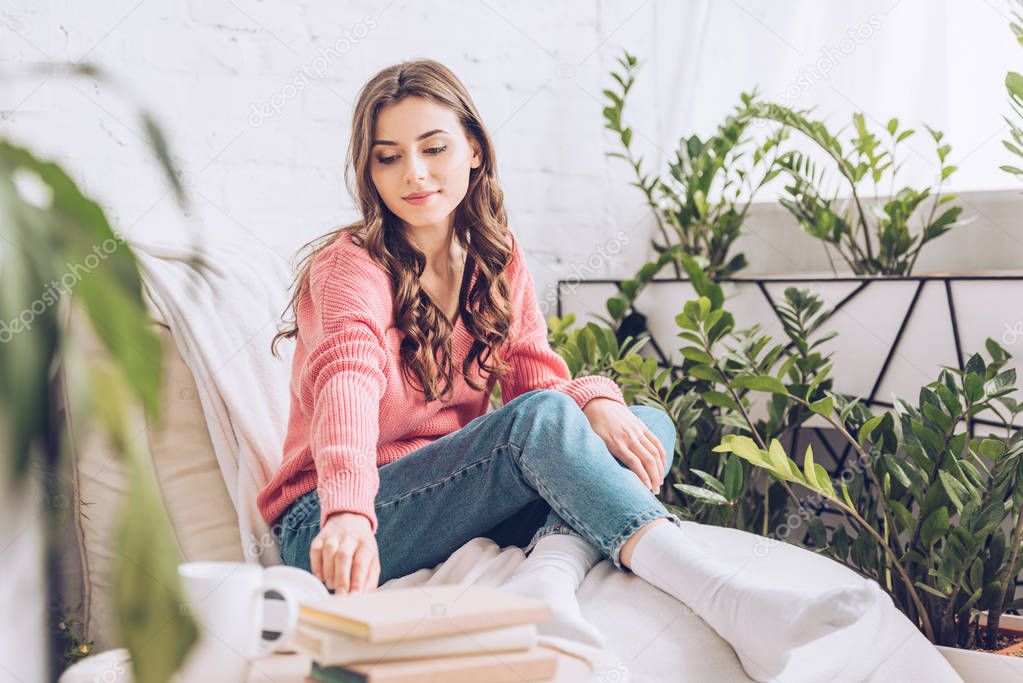 selective focus of attractive girl taking book while sitting in white soft chaise lounge at home