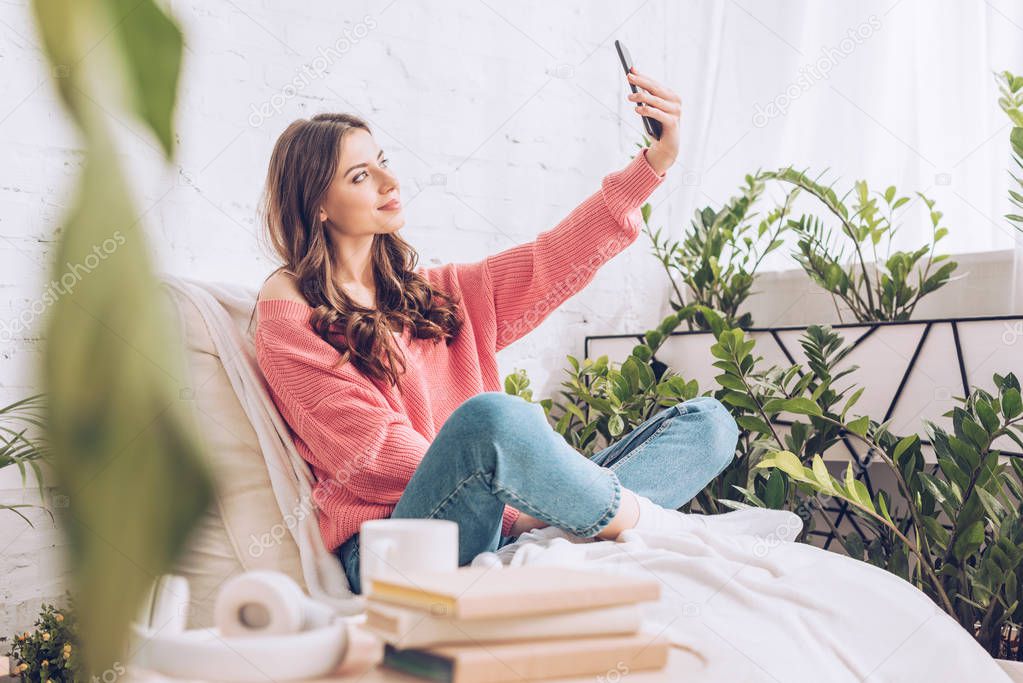 selective focus of smiling young girl taking selfie with smartphone while sitting with crossed legs on soft chaise lounge