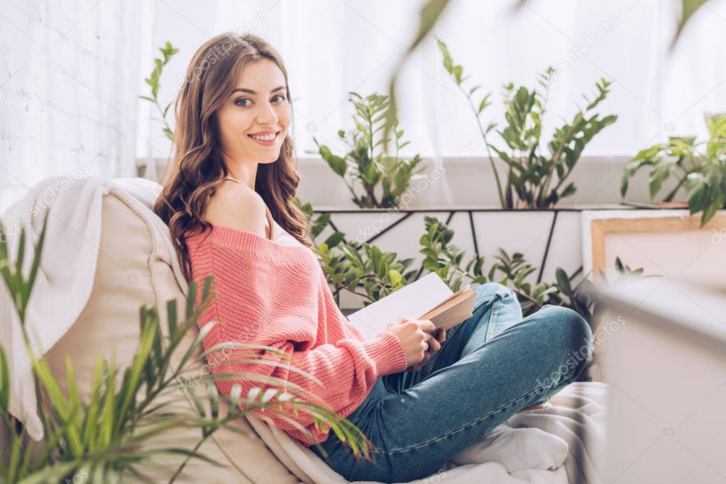 selective focus of cheerful girl holding book and smiling at camera while sitting in room with green plants 