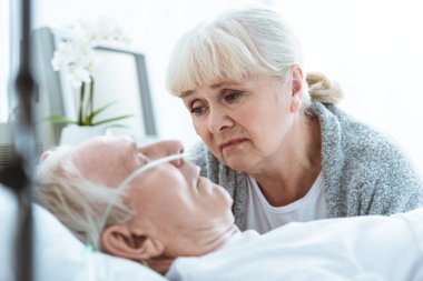 sad senior woman looking at husband in coma in clinic clipart
