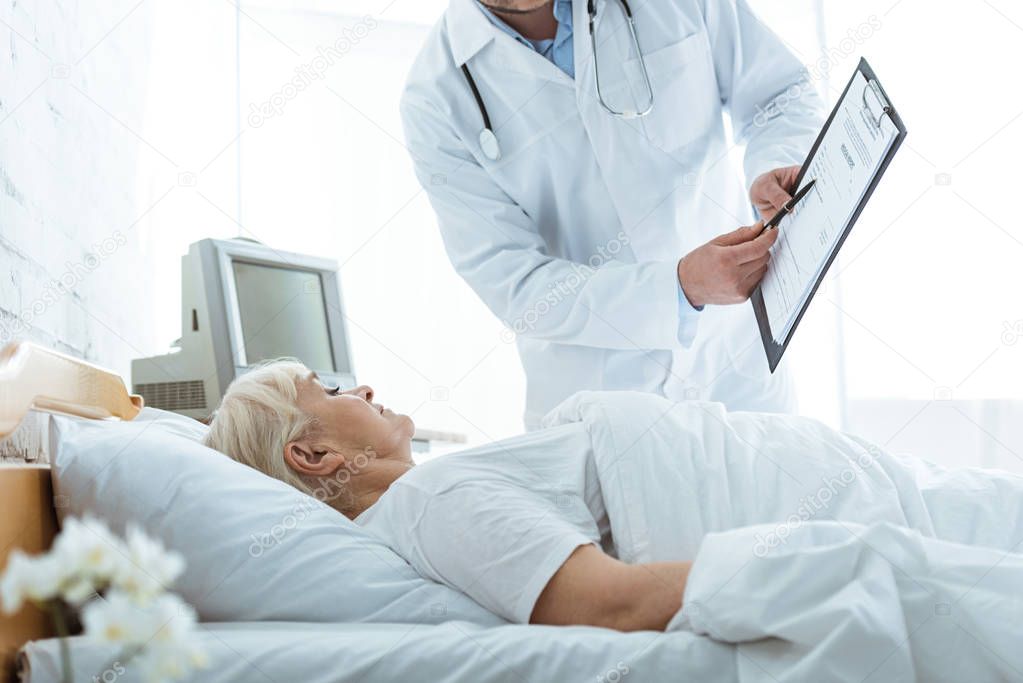 cropped view of doctor with clipboard and sick senior woman lying on bed