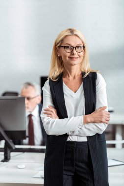 selective focus of cheerful blonde businesswoman in glasses standing with crossed arms near coworker  clipart