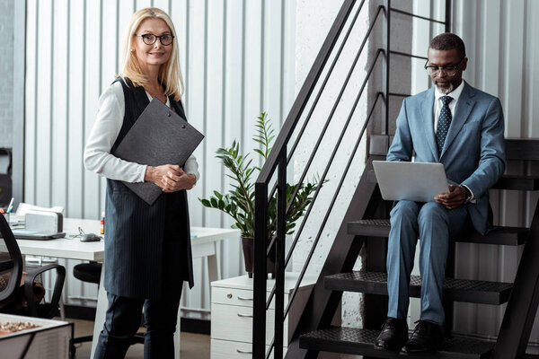 cheerful blonde woman with clipboard and standing near african american man using laptop 