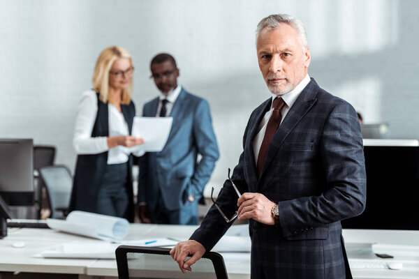 избирательный фокус бизнесмена, держащего очки и смотрящего в камеру, стоя рядом с коллегами
 