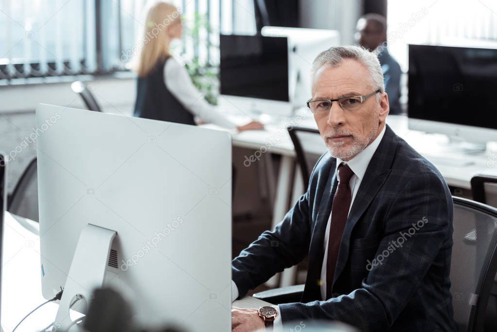 selective focus of mature businessman in glasses looking at camera near multicultural coworkers 