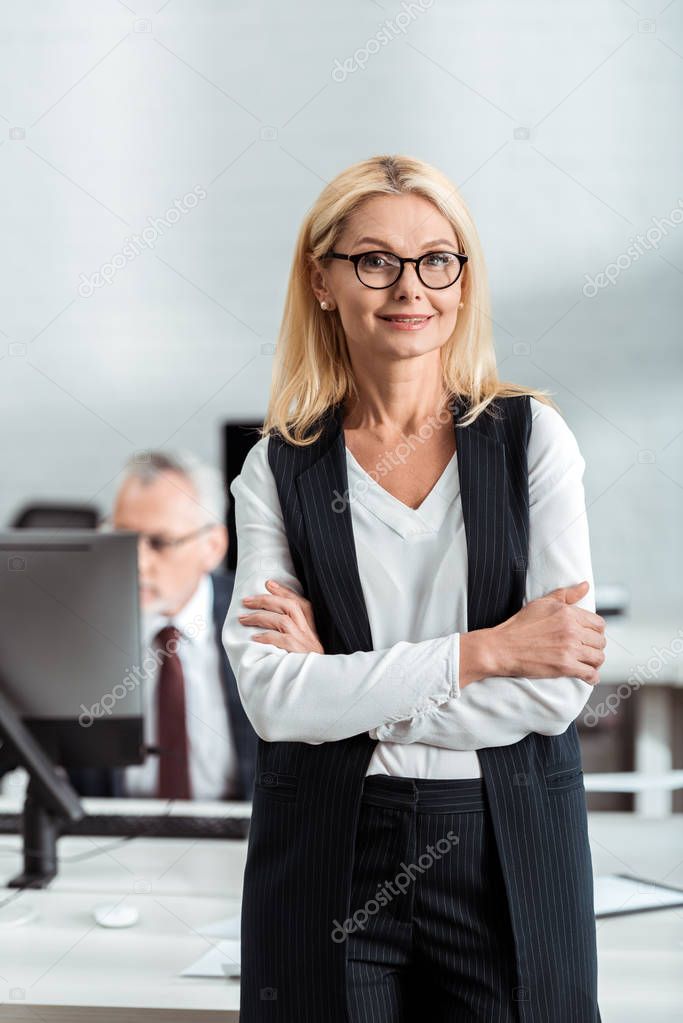 selective focus of cheerful blonde businesswoman in glasses standing with crossed arms near coworker 