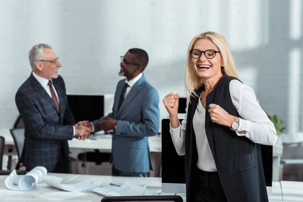 多文化ビジネスマンがオフィスで握手をする近くで勝利を祝う幸せなビジネスウーマンの選択的な焦点 — ストック写真