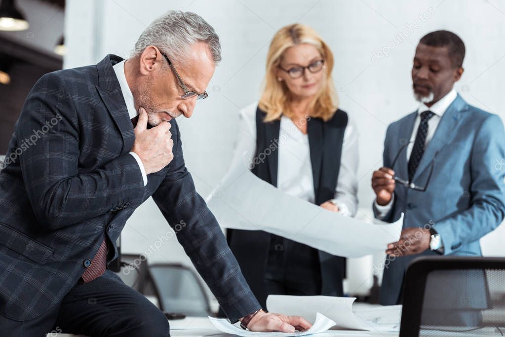 selective focus of thoughtful businessman standing near multicultural partners with blueprints 