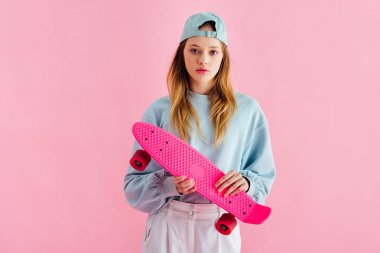 pretty teenage girl in cap holding penny board isolated on pink clipart