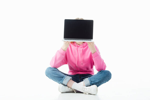 teenage girl with obscure face and crossed legs holding laptop with blank screen isolated on white, illustrative editorial