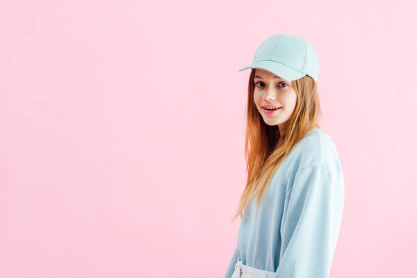 happy pretty teenage girl in cap looking at camera isolated on pink