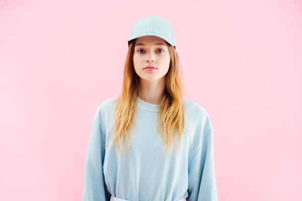 sad pretty teenage girl in cap looking at camera isolated on pink