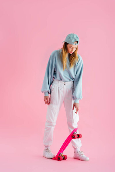 pretty teenage girl in cap riding penny board on pink