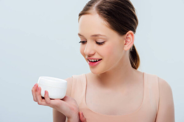 cheerful teenage girl looking at cosmetic cream isolated on grey
