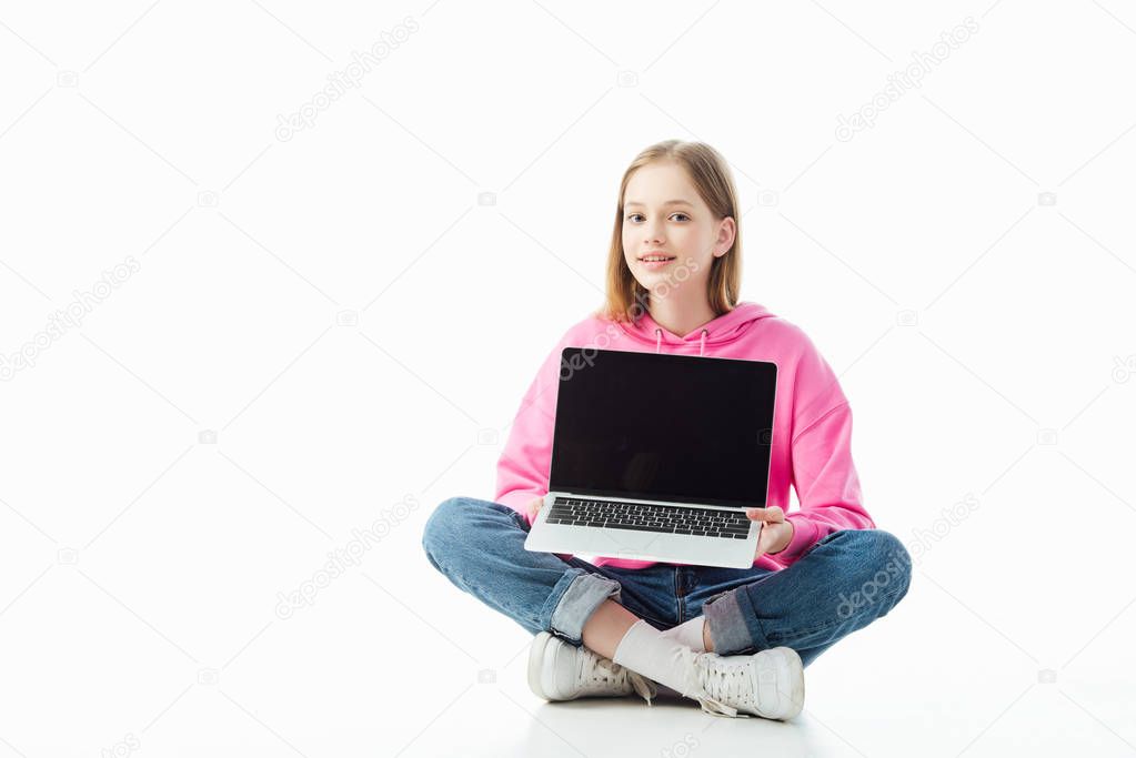 smiling teenage girl with crossed legs holding laptop with blank screen isolated on white, illustrative editorial