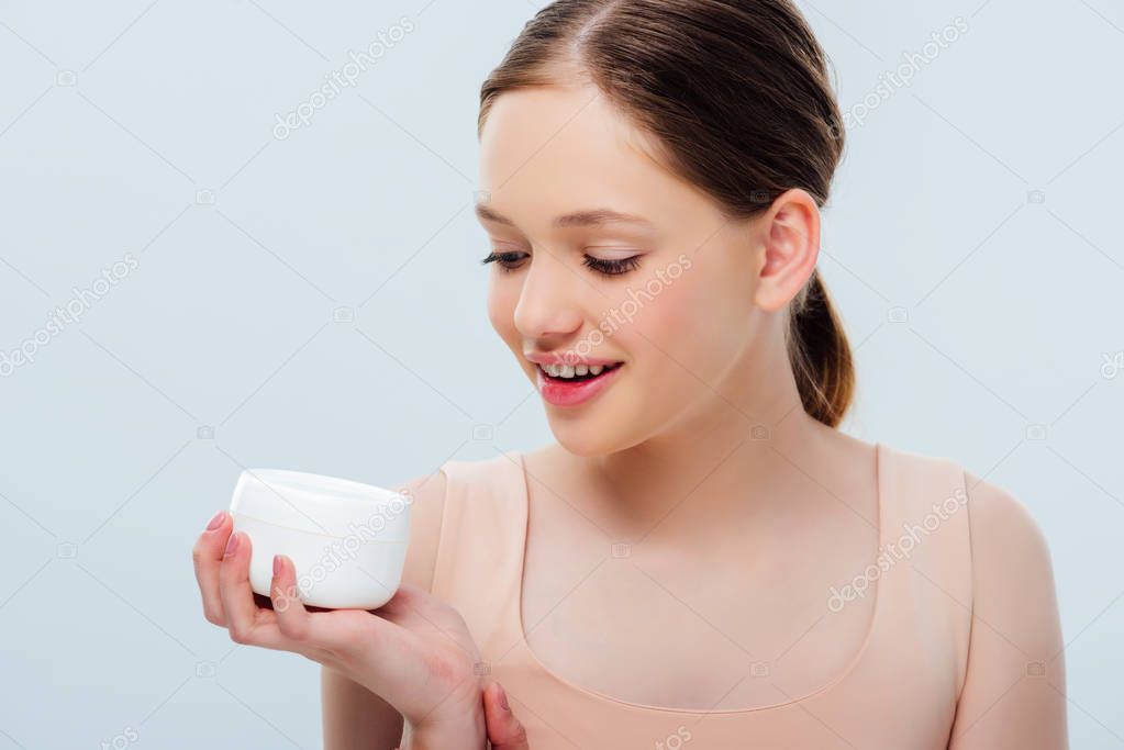 Cheerful teenage girl looking at cosmetic cream isolated on grey