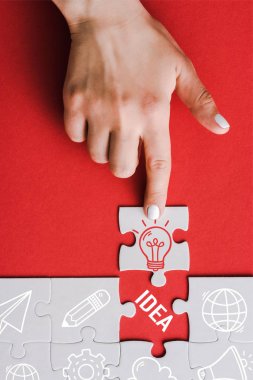 top view of woman pointing with finger at jigsaw with light bulb near connected white puzzle pieces and idea lettering on red clipart