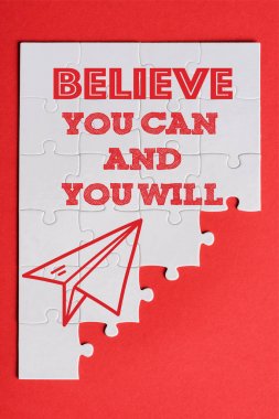 top view of white connected puzzle pieces with paper plane and believe you can and you will lettering isolated on red  clipart
