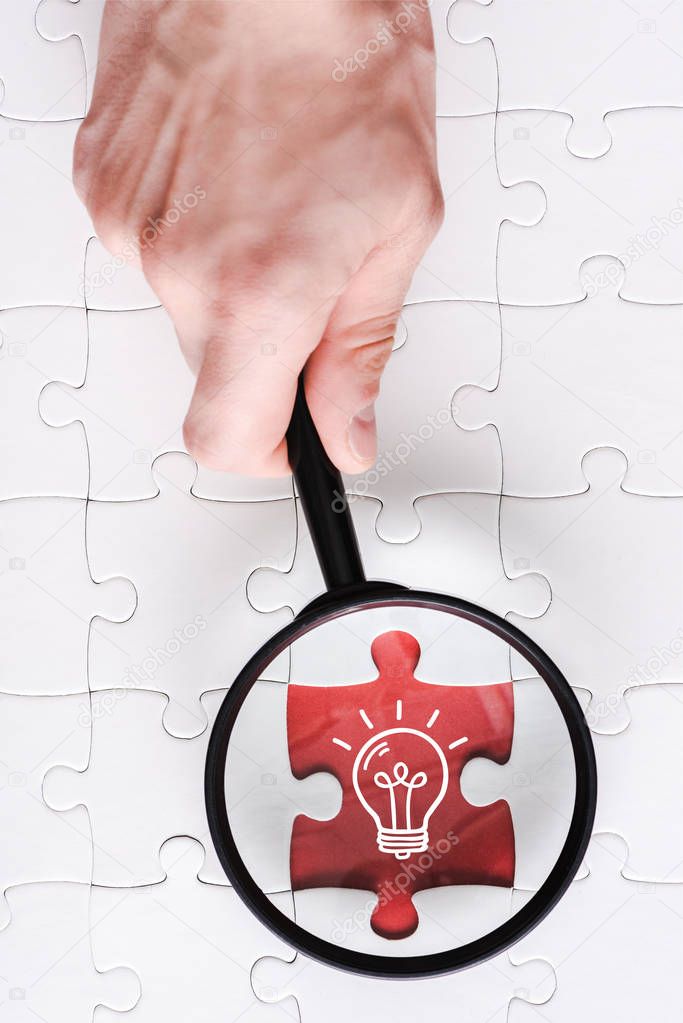 top view of man holding magnifying glass near jigsaw with light bulb near white connected puzzle pieces 