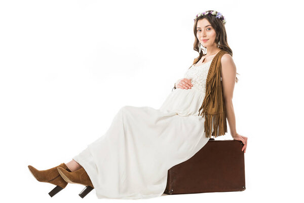 Улыбающаяся беременная женщина в венке сидит на чемодане изолированы на белом
