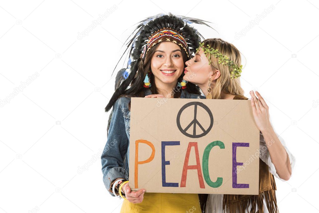 two bisexual hippie girls in indian headdress and wreath holding placard with inscription and kissing isolated on white