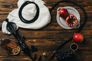 top view of hookah, cinnamon, garnets, grapes and dried oranges on wooden surface clipart