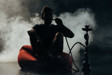 silhouette of man sitting on bean bag chair and smoking hookah in darkness clipart