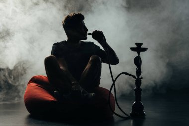 silhouette of man sitting on bean bag chair  with crossed legs and smoking hookah clipart