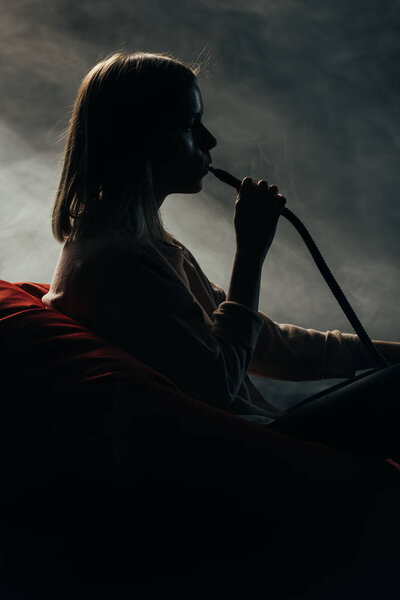 silhouette of woman sitting on bean bag chair and smoking hookah in darkness