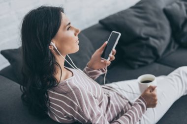 boş ekranlı akıllı telefon tutan ve kulaklıkla müzik dinlerken uzağa bakan düşünceli genç kadın