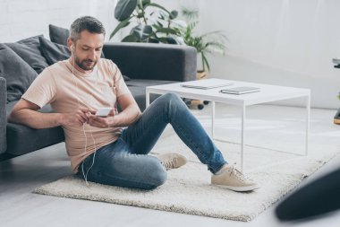 yakışıklı adam evde katta oturan, akıllı telefon ve kulaklık müzik dinleme kullanarak