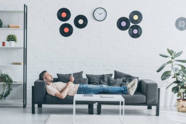 adult man in earphones using smartphone while lying on sofa in spacious living room clipart