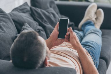kulaklıklı adamın seçici odak kanepede yatan ve boş ekranlı akıllı telefonu tutan