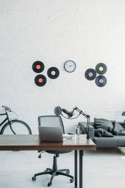 ahşap masa, ofis sandalyesi, vinil diskler ve beyaz duvarda saat mikrofon ve dizüstü bilgisayar ile geniş yayın stüdyosu