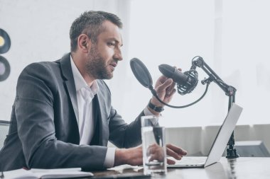 yakışıklı radyo host mikrofonda konuşurken dizüstü bilgisayar ve su bardağı yakınında işyerinde otururken