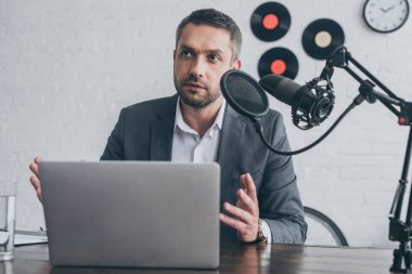 handsome radio host gesturing while speaking in microphone at workplace near laptop  clipart
