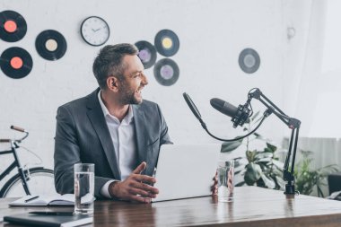 mikrofon ve dizüstü bilgisayarın yanında masada otururken gülen neşeli radyo host